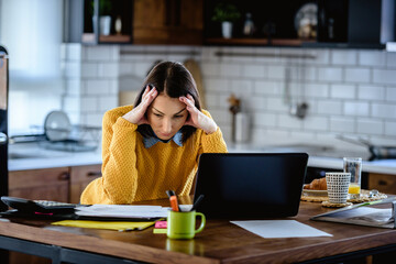 Worried businesswoman working online from home in front of a laptop computer looking into papers....