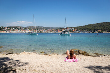 Girl lying on the beach while relaxing and sunbathing, watching anchored sailing boats in the bay of Rogoznica, Croatia