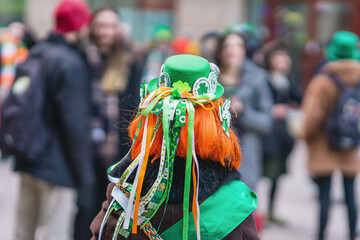 Back view of girl with red hair in hat with decorations, symbols of St. Patrick's Day, parade in...