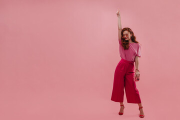 In full growth, confident caucasian young lady raises hand straight up while in photo studio. With loose red hair, eyes closed, is standing, dressed in T-shirt, loose pants and sandals with heels.