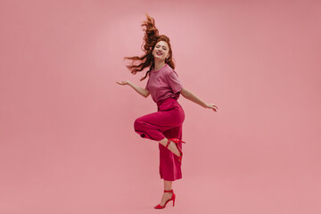 In full growth, fantastic caucasian young girl stands sideways with leg curled up at knee on pink studio background. Redhead model waving long hair smiling at camera, wearing t-shirt, pants and heels.