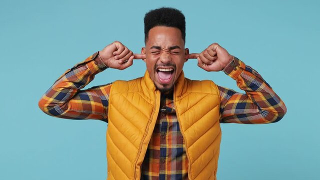 Displeased irritated sad young african american†man 20s wears yellow shirt waistcoat closed eyes cover ears do not want to listen scream isolated on plain pastel light blue background studio portrait