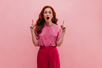 Caucasian young girl shows index fingers upward on pink background with place for text. Long-haired...