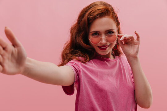 Close-up of amazing caucasian young girl makes photo with glasses on her face on pink background. Fair-skinned redhead lady is looking at camera, wearing T-shirt. Free time for selfies.