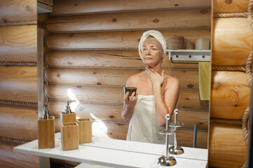 A woman takes care of herself standing in the bathroom of a wooden house. Eco-friendly self-care. Natural cosmetics. A woman applies a natural brown coffee mask to her face and neck.