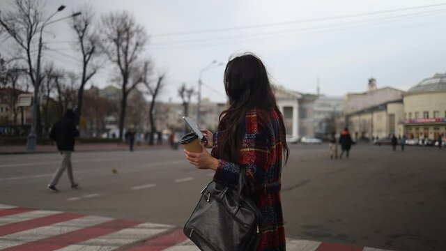 Asian woman in coat with smartphone walking against city street.  Fashion business photo of beautiful girl in red suit with smart phone and coffee.