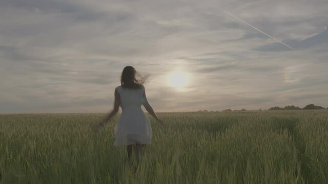 A young beautiful girl in a white dress runs along a green wheat field at sunset