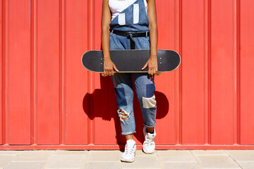 Unrecognizable woman holding a skateboard with her hands on a red urban wall.