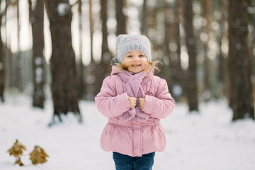 Winter in the forest. Cute dreamy little girl in a pink coat standing in snowy forest with trees background and looking on camera. Smiling kid walking in frosty winter day in forest.