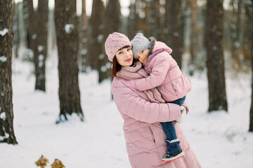 Family on a walk in winter forest. Young mother hugging and smiling with her little daughter on hands in beautiful winter snowy forest. Happy motherhood concept.