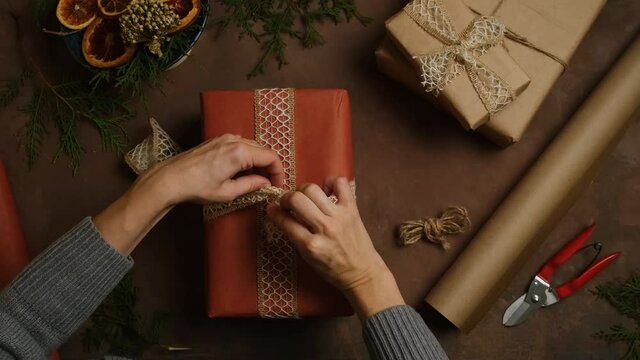 Woman decorates Christmas present, tie a bow on red gift box. Female hands packages Christmas gift. Overhead view.