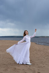 Fototapeta na wymiar Happy young girl in a white dress on the river bank. Stormy sky over the river