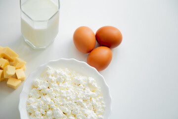 A white bowl with cottage cheese, eggs, diced cheese and a glass of milk stand on a white background. . High quality photo