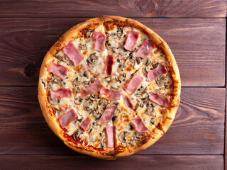 Close-up pizza with ham and mushrooms on a brown wooden background. A traditional Italian dish. A delicious and unhealthy dish. Top view, flat lay