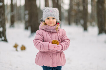Portrait of little child girl, wearing pink warm coat and gray cap, posing on fresh air in winter snowy forest with tasty homemade cookie in hands.