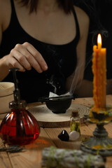Person throwing herbs in a small cauldron, where a paper burns, smoke coming out of it. It sits on a wooden surface on which there are more witchy items like candles, cristals and a mortar.