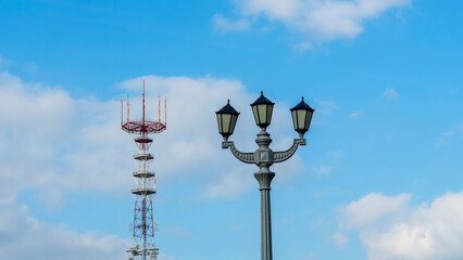 A TV tower on the background of a blue sky with clouds. In the foreground is a vintage street lamp.