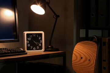 Clock with glowing lamp and computer on table in dark room