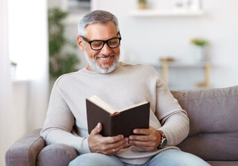 Happy senior man with grey hair in eyeglasses reading book at home, resting in living room
