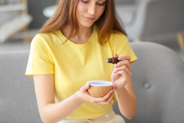 Young woman sitting on sofa and adding essential oil to diffuser, closeup