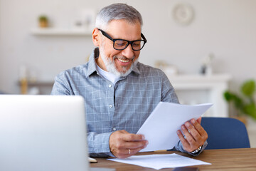 Handsome smiling senior man reading financial documents while working remotely on laptop at home