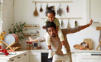 Cute little african american child son sitting on father shoulders while playing together at home