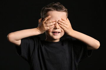 Portrait of cute little boy covering eyes with his hands on dark background