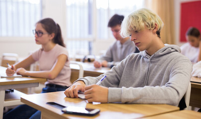 Fototapeta premium Smart teenager studying in classroom, listening to lecturer and writing in notebook..