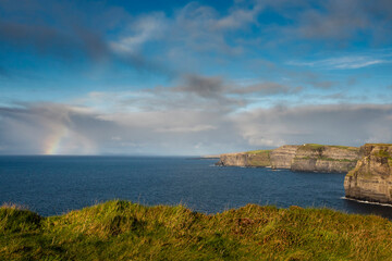 Naklejka premium Colorful rainbow in the ocean next to Cliff of Moher, county Clare, Ireland. Amazing scenery with great view. Cloudy sky and blue ocean water. Top Irish nature landmark and travel destination