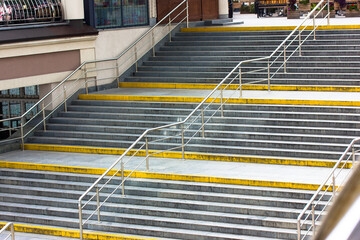 A wide, modern stone staircase with yellow markings for blind people and metal handrails for a...
