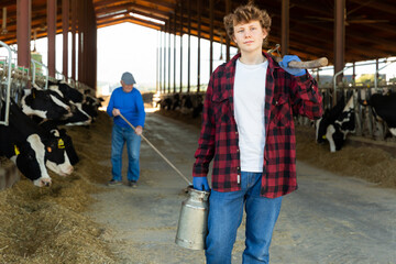 Confident teenage farm worker walking through cowshed after work on background of stall with cows,...