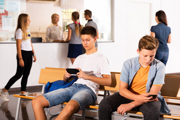 Two teenagers using smartphones sitting on bench in college hall