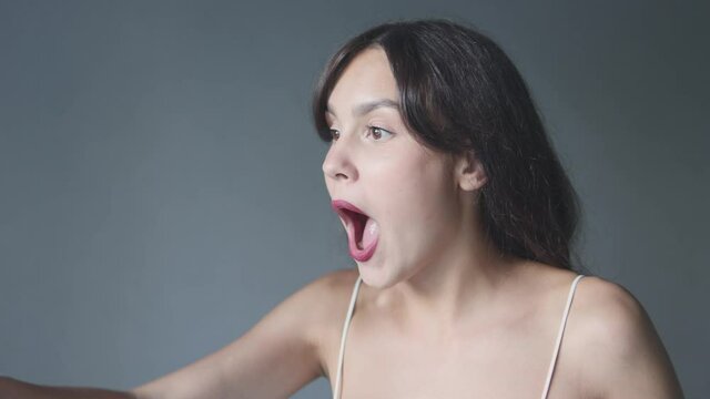 expressive caucasian brunette female 20 saying wow wondering on grey background copy space. portrait young woman chocked surprised mouth opened hands cheeks big eyes wide. emotions advertising concept