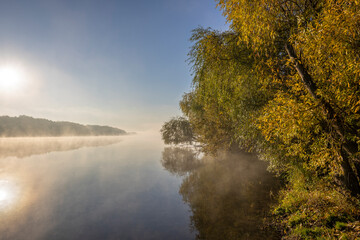 Naklejka na ściany i meble Autumn landscape in the early morning on the river. Misty water surface. Yellow leaves on trees and bushes are illuminated by the rays of the rising sun. Dawn on a cold autumn morning.