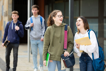 Young cheerful teenager girls with workbooks in hands laughing outdoors near college building