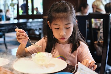 A cute young Asian girl is sitting in a restaurant, eating a bowl of vanilla ice cream, decorated by white smoke from dry ice, with cherry topping.  Kid feeling happy to eat ice cream dessert.