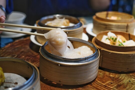 A close-up picture of bamboo chopsticks picking up white white Chinese stuffed steamed dumpling, also known as dim sum from a round bamboo basket on a nice dinner table.
