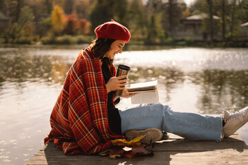 A Teengirl in a red beret reading book on wooden pontoon. Autumn season. Female with trendy clothes...