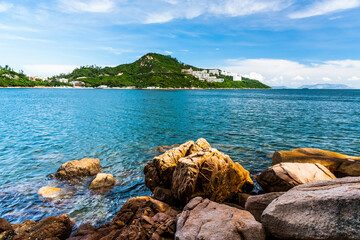 The rocky seashore of Stanley Bay on Hong Kong Island in Hong Kong. Beautiful scenic landscape with water, mountains, rocks, buildings, fishing boats, and cloudy sky.