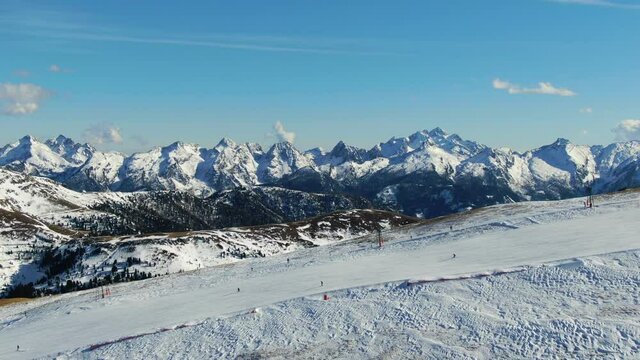 Aerial drone shot with a parallax effect of ski riders going down the hill with beautiful peaks of the tyrol mountains in the back on a sunny day during ski season.
