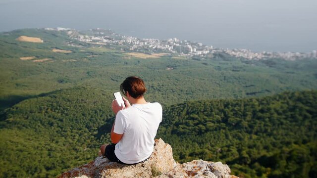 Adventure trip - young man hiker sitting on the edge of a rock and takes photos of the forest and mountains on his phone