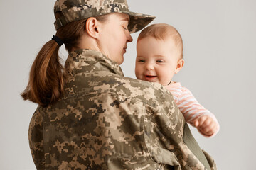 Back view of woman soldier wearing camouflage uniform and cap, hugging her infant daughter while holding her in hands, returning home from army or war.