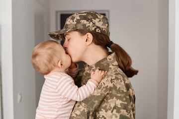 Lovely young adult woman soldier wearing camouflage uniform, returning home and hugging her little...