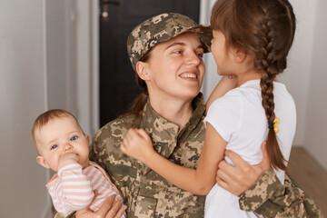 Closeup portrait of happy military mother wearing camouflage uniform and hat, hugging her daughters while returning home after served in army, expressing happiness.
