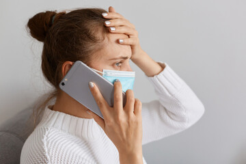 Side view of unhealthy woman wearing white sweater and medical protective mask talking mobile...