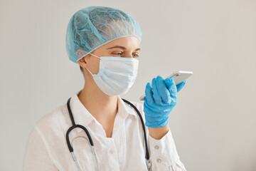 Indoor shot of woman doctor wearing medical cap, gloves and protective mask using mobile phone,...