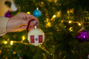 Close up of girls hand holding a Christmas ball for a fir tree with the flag of Peru. New year in Peru. Christmas holiday greeting card with copy space for text. Decorating home for celebration.