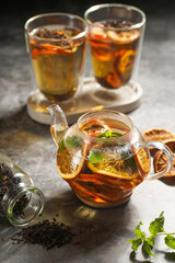 A transparent tea pot filled with indian black tea, oranges and mint on a grey surface