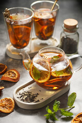 A transparent tea pot filled with black tea, oranges and mint on a grey surface