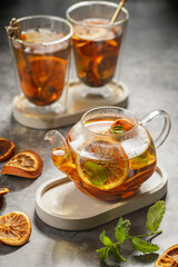 A transparent tea pot filled with black tea, oranges and mint on a grey surface